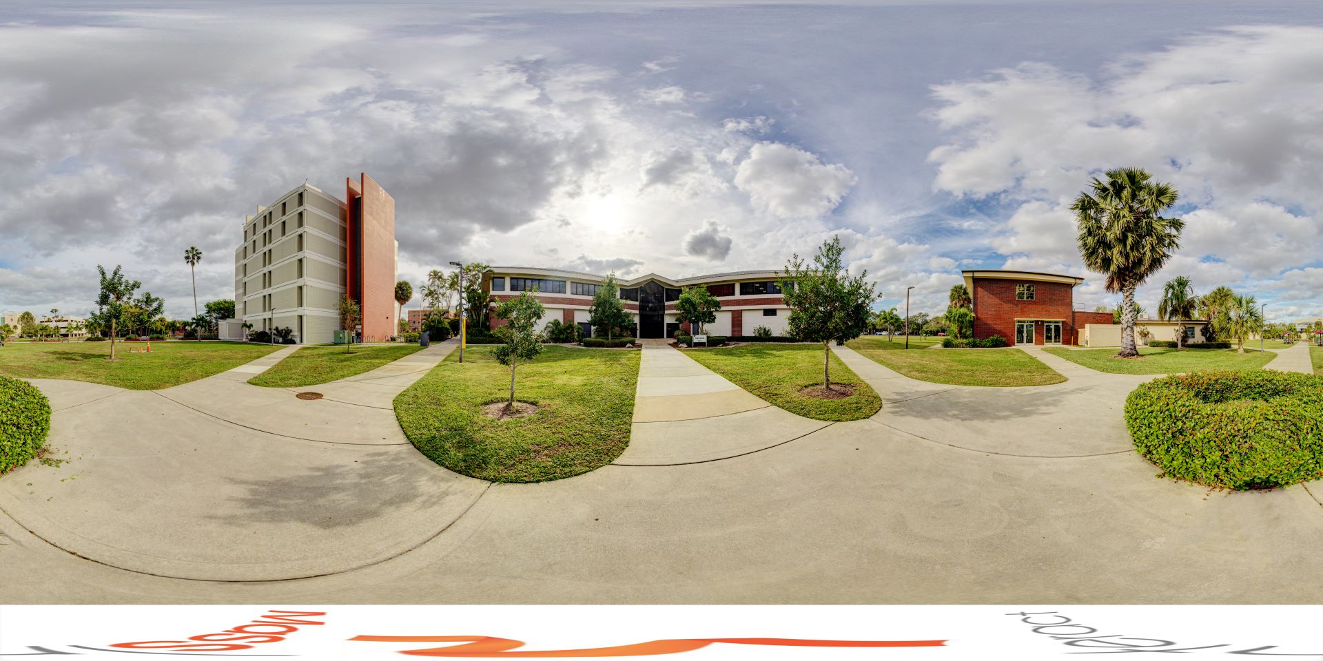 Panoramic view of a university campus with Skurla Hall, a tall white building with an orange stripe, surrounded by green lawns, walking paths, and palm trees under a partly cloudy sky